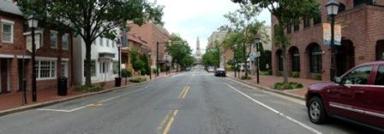 This photo shows 1 of 14 roadway panoramas presented to participants in the similarity rating task. There is a straight two-lane roadway with on-street parking on both sides of the roadway, although only two parked cars are visible. There are red-brick sidewalks on both sides of the roadway. Red-brick buildings of two or three stories abut the sidewalk. There are decorative street lamps and planters at intervals on the sidewalks on both sides. Directly ahead, in the far distance, an obelisk is aligned with the center lane marking.