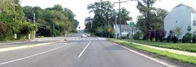 This photo shows 1 of 14 roadway panoramas presented to participants in the similarity rating task. A four-lane undivided roadway curves to the left. The asphalt has many cracks. A sidewalk abuts the roadway on both sides. A driveway on the right provides access to a gas station and garage. On the left side of the road, a parking lot is visible. There is a restaurant at the far end of that lot. A speed limit sign is attached to a power pole that is in the sidewalk on the right side of the roadway.