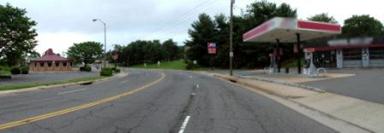 This photo shows 1 of 14 roadway panoramas presented to participants in the similarity rating task. A four-lane undivided roadway curves to the left. The asphalt has many cracks. A sidewalk abuts the roadway on both sides. A driveway on the right provides access to a gas station and garage. On the left side of the road, a parking lot is visible. There is a restaurant at the far end of that lot. A speed limit sign is attached to a power pole that is in the sidewalk on the right side of the roadway.