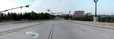 This photo shows 1 of 14 roadway panoramas presented to participants in the similarity rating task. A straight four-lane divided roadway with an additional left-turn lane is shown. A signalized intersection is ahead. A concrete barrier to the right of the roadway separates the road from a sidewalk that is not visible. There is a metal railing above the concrete barrier. At intervals, there are decorative street lamps on poles attached to the concrete barrier. In the distance, on the right, there is a high-rise apartment building and a large water tank. Trees are visible ahead on the left.