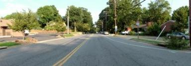 This photo shows 1 of 14 roadway panoramas presented to participants in the similarity rating task. A two-lane roadway with a double yellow centerline appears to be entering a residential neighborhood. On the right is a sidewalk, which is separated from the roadway by a thin grass strip. Wooden power poles rise from the thin grass strip. A parking lot and commercial building are to the left of the roadway. A speed limit sign is attached to a power pole on the right. In the distance, trees line both sides of the road.