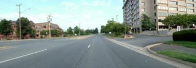 This photo shows 1 of 14 roadway panoramas presented to participants in the similarity rating task. A four-lane roadway through a commercial area is shown. A wide driveway on the right provides access to a mid-rise concrete and glass building. There is an asphalt sidewalk that is separated from the roadway by a thin grass strip. There are brick buildings on the left side of the roadway. The landscape includes a few small trees on both sides of the road. Power poles are visible on the left side of the road.