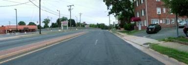 This photo shows 1 of 14 roadway panoramas presented to participants in the similarity rating task. A straight four-lane road appears to curve to the right in the distance. The road is just wide enough for the four lanes and a small brick-lined median. There are sidewalks on either side, which are separated from the road by a thin grass strip. Ahead on the right is a parking lot and older red-brick building. On the left is a larger parking lot and what appears to be a store set back from the road. Power lines are prominent on the left side of the road.