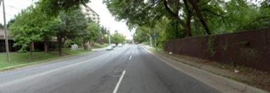 This photo shows 1 of 14 roadway panoramas presented to participants in the similarity rating task. Travel lanes abut the curb and leave no room for parking. An unimproved dirt path abuts the curb on the right. A wooden fence, more than 6 ft (2 m) high, is to the right of the dirt path. Trees lean over the fence and shade the road. On the left side of the road, a mowed lawn slopes upward away from the curb. There are a few trees on the lawn. At the top of the lawn is another fence that looks similar to the one on the right.
