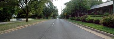 This photo shows 1 of 14 roadway panoramas presented to participants in the similarity rating task. A straight four-lane divided road is shown. Travel lanes abut the curb and leave no room for parking. The grassy median has trees at intervals. No sidewalks are visible. A neatly trimmed grass strip, about 3 ft (0.9 m) wide, abuts the curb on the right side. Beyond the grass strip is a row of neatly trimmed bushes. Behind the row of bushes is more grass and a single-story building with what appears to be a metal roof. To the left side of the road are trees and a white painted wall that frames both sides of a driveway. In the distance, trees line both sides of the road. A yellow warning sign is visible ahead on the right side of the road.