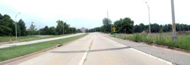 This photo shows 1 of 14 roadway panoramas presented to participants in the similarity rating task. A straight four-lane divided highway is shown. The travel-way is concrete, and the shoulder is asphalt. Weeds grow along the curb that abuts the asphalt shoulder. The median has mown grass, but no trees or other plants. Street lights of the type used on freeways and other major roads are on both sides of the highway. There is a yellow warning sign ahead on the right. Trees are visible in the distance, but the nearby areas are covered by either grass or tall weeds.