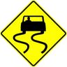 The slippery when wet symbol sign is shown. The warning sign is a yellow diamond, and the symbol consists of a silhouette of the rear of a passenger car with S-shaped curves trailing each rear tire.