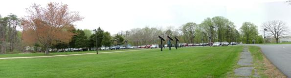 A panoramic photograph is shown. An area with a green lawn in the foreground is vertically bisected by a walkway. A parking lot full of cars can be seen beyond the lawn and walkway.