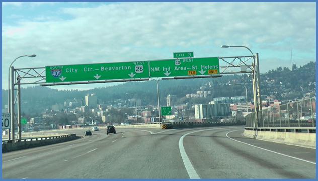 This photo shows the driving image for scenario 1 critical point 2. There is a four-lane single direction highway. Two lanes split to the right, and three lanes split to the left. The second-to-right lane is an option lane. There is a sign that spans the three lanes splitting to the left. Each of those three lanes has a down arrow with the words  City Ctr.-Beaverton  above the arrows. The two lanes that split to the right each have a down arrow and the words  N.W. Ind. Area - St. Helens  above the arrows. There is a yellow sign indicating that the rightmost lane is an exit only lane. There is also a sign above both of these lanes labeled  Exit 3. 