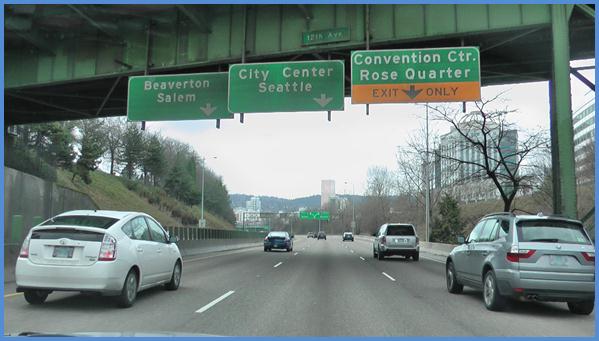 This photo shows the driving image for scenario 2 critical point 1. There is a three-lane single direction highway with cars traveling on it. There are three green signs: one above each of the lanes. The sign on the left has a down arrow and is labeled  Beaverton Salem.  The middle sign has a down arrow and is labeled  City Center Seattle.  The sign on the right is green on the top half and is labeled  Convention Ctr. Rose Quarter.  The bottom half is yellow with a down arrow and is labeled  Exit Only. 