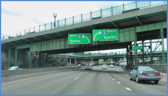 This photo shows a driving image for scenario 3 critical point 3. There is a three-lane single direction highway with cars traveling on it. There are two green signs: the one on the left spans the left lane and the left half of the middle lane, and the sign on the right spans the right half of the middle lane and the right lane. The sign on the left shows an arrow curving to the left and is labeled  5 South Salem.  The sign on the right shows an arrow curving to the right and is labeled  5 North Seattle. 