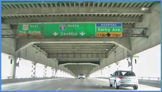 This photo shows a driving image for scenario 3 critical point 1. There is a four-lane single direction freeway with vehicles traveling on it. There are three signs spanning over the four lanes. The sign on the left is directly over the left-most lane. The top half is green and is labeled  30 East The Dalles.  The bottom half is yellow with a down arrow and is labeled  Exit Only.  The middle sign is green and spans over the two middle lanes. There are two down arrows, and it is labeled  5 North Seattle.  The right-most sign is directly over the right-hand lane. The top part is blue and is labeled  Hospital.  The middle part is green and is labeled  Kerby Ave.  The bottom part is yellow with a down arrow and is labeled  Exit Only. 