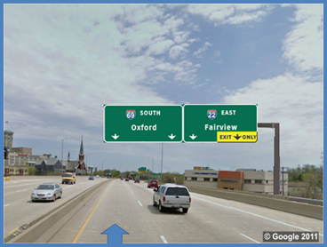 This photo shows an example test slide from topic 1 depicting a through lane to exit movement. There is a three-lane single direction highway with vehicles traveling on it. There is a blue arrow pointing up in the left lane. There are two signs over the three lanes. The sign on the left has two arrows: one pointing to the left lane and the second pointing to the middle lane. Above the arrows, the sign is labeled  69 South Oxford.  The sign on the right also has two arrows: one pointing to the middle lane and the second pointing to the right lane and labeled  Exit Only  with a yellow background. Above the arrows, the sign is labeled  22 East Fairview. 