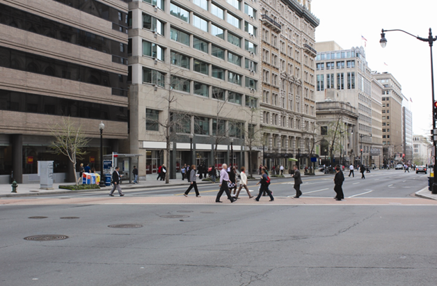 An image of the southernmost crossing of 14th Street Northwest at New York Avenue Northwest. The photo is taken from the northwest corner of the intersection. There are approximately nine people crossing in the marked crosswalk and two people crossing between the marked crosswalks.