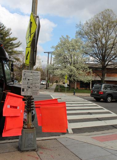 An image of the (far) flag crossing at Connecticut Avenue Northwest and Northampton Street Northwest. A pole with several orange flags and an instruction placard can be seen in front of the marked crossing.