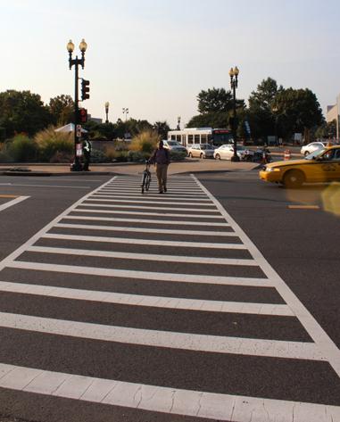 Illustration of what a pedestrian might see as he or she attempts to cross from the north side of Independence Avenue Southwest to the south side of the street along Washington Avenue Southwest. The photo is taken from the pedestrian perspective. A pedestrian can be seen walking a bicycle across the roadway in the marked intersection. A left-turning taxi cab can be seen waiting for the pedestrian to complete the crossing prior to passing through the crosswalk.