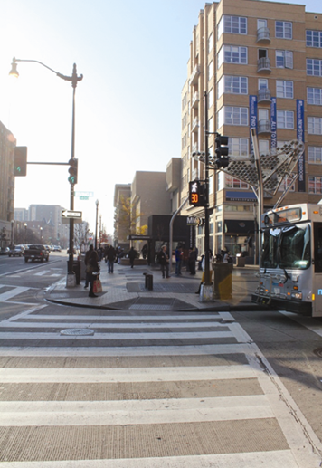 Illustration of what a pedestrian might see as he or she attempts to cross from the north side to the south side of Irving Street Northwest along 14th Street Northwest in Washington, DC. Pedestrians can be seen across the street congregating by a Metro station. The photograph is taken from the pedestrian perspective.