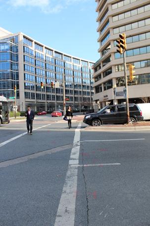 Illustration of what a pedestrian might see as he or she attempts to cross from the west side to the east side of Wisconsin Avenue along Willard Avenue in Chevy Chase, MD. Cross traffic can be seen stopped behind the marked crosswalk while two people are crossing. The photograph is taken from the pedestrian perspective. 