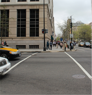 This photo shows a typical crosswalk in Washington, DC. Two straight, parallel lines connect two street corners, leaving enough room in between the lines for a large group of pedestrians to cross the street. Two cars have stopped behind one of the lines before entering the intersection, illustrating how drivers yield when approaching a crosswalk.