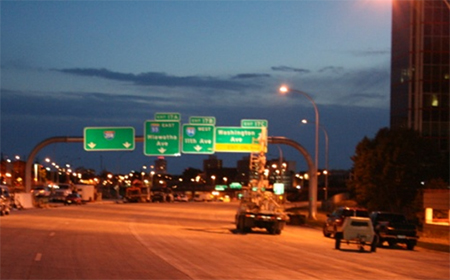 Figure 2. Photo. Roadway lighting. Figure 2 is a photograph of a stretch of freeway in a congested area. In the photograph, typical highway signs are present, as are overhead luminaires. To the right of the photo is a tall building and to the left are a number of construction vehicles parked on the shoulder.