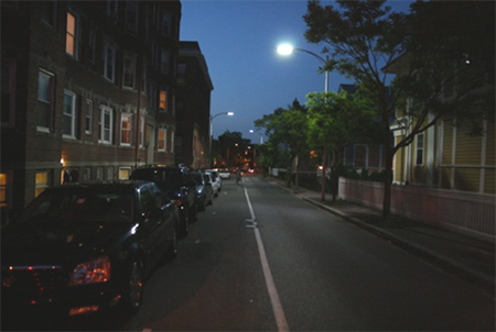 Figure 3. Photo. Street lighting. Figure 3 is a photograph of a stretch of urban local road. On the left are row houses and parked cars, and on the right are a sidewalk, trees, and individual houses. Overhead luminaires are on both sides of the road.