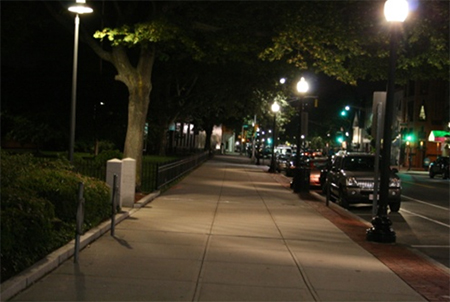 Figure 4. Photo. Residential/pedestrian area lighting. Figure 4 is a photograph of an urban or suburban sidewalk. On the left is green space and on the right is the road, with parked cars and storefronts. Post-top luminaires line the roadway side of the sidewalk, and one small overhead luminaire is on the left of the sidewalk in the green space.