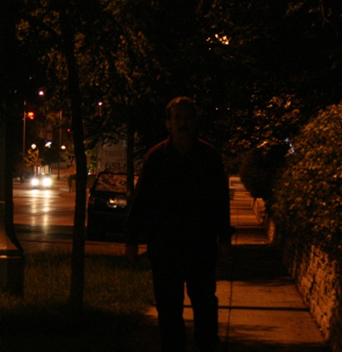 Figure 6. Photo. Facial recognition under low lighting. This image features a roadway on the left and a brick retaining wall on the right. In the approximate center of the photo is a male pedestrian. Overhead luminaires are not illuminated, there is low lighting, and the man’s face is not recognizable.