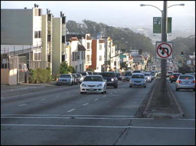 Figure 10. Photo. Example of an urban arterial in a residential area. A street-level photo shows an example of an urban arterial in a residential area. The arterial is a six-lane cross-section divided by a narrow, raised median. There are apartment buildings along the left side of the corridor, and the other side is not visible in the photo. There is lighting installed along the raised median and a “no left-turn or u-turn” sign installed on the nearest light pole. Cars are visible in all travel lanes and parked on-street in front of the apartment buildings.