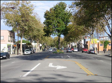 Figure 11. Photo. Example of a suburban arterial in a commercial area. A street-level photo shows an example of a suburban arterial in a commercial area. There are small business buildings along both sides of the corridor. The arterial is a four-lane cross-section divided by an intermittent raised median. Where there is no raised median, there are left-turn lanes. Cars are visible in the travel lanes and parked along both sides of the street.