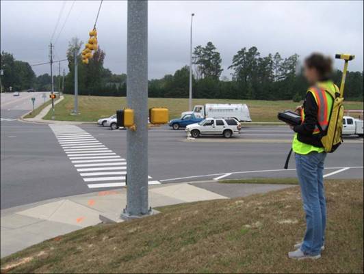 Figure 16. Photo. Data collection equipment used for field visit. A photograph shows a data collector standing at the corner of a signalized intersection, holding a tablet PC and noting specific characteristics of the intersection, while the GPS device is mounted atop a backpack system.