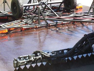 The photo shows a brown liquid being sprayed from  nozzles, leaving a shiny brown coating on the pavement surface. A device with  wheels and brushes towed behind the nozzles scrubs the liquid into the surface.