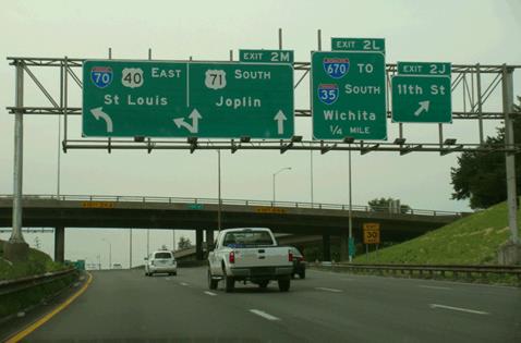 This photo shows vehicles on a highway. The highway has four direction signs. One sign has no direction arrow, but the other three signs do have direction arrows. On the signs that have upward-facing arrows, the length of the arrows is truncated, reducing the height of the sign.