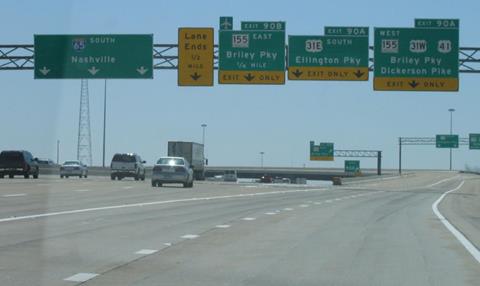 This photo shows vehicles on a highway. Five signs hang above the highway. The signs are placed very closely together and have so many directions, destination names, and arrows on them that a driver is liable to become confused.