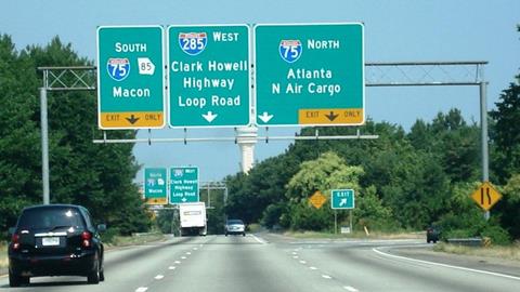 The photo shows vehicles on a highway with a right-hand exit ramp. Three signs with directional arrows and destination names hang above the highway. The signs are not directly hanging over the lanes that their arrows are meant to point to.