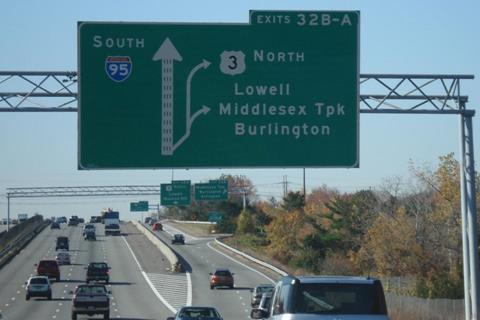 This photo shows vehicles on a highway. The right lane of the highway branches off into a separate lane (separated from the highway by a concrete median) that has its own right-hand exit ramp. A direction sign hangs above the highway and illustrates both the beginning of the separate lane and the exit ramps off of that lane.