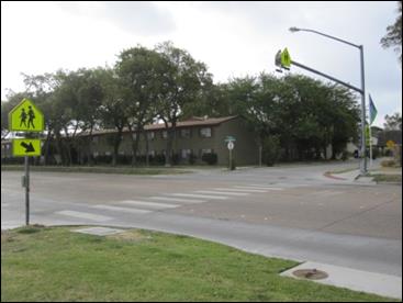 Figure 1. Photo. School crosswalk with RRFBs in Garland, Texas. A pedestrian crosswalk in a school zone. The crosswalk is supplemented by rectangular rapid flashing beacons, one set of post-mounted beacons on each side of the road, and one set above the center of the road on a mast arm.