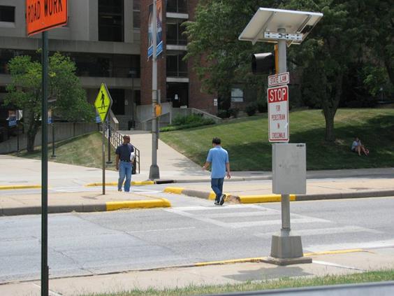 Figure 13. Photo. Unsignalized pedestrian crosswalk in Kansas City, KS. The midblock crosswalk is marked with a ladder design and crosses a roadway with four through lanes and a raised median. The crosswalk cuts through the raised median, and there is a pedestrian crosswalk sign (W11-2) located in the raised median. There is a white and red sign on the right side of the roadway that reads “State Law: Stop for Pedestrian in Crosswalk.” There are two pedestrians crossing the roadway—one in each direction.