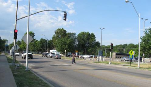 Figure 14. Photo. Signalized pedestrian crosswalk in Kansas City, MO. The midblock crosswalk is marked with a ladder design (although the painted marking is worn) and crosses a roadway with four through lanes and a two-way left-turn lane. Parking is allowed on one side of the roadway. There is a pedestrian crossing the roadway. The signal is red. There is a pedestrian crosswalk sign (W11-2) at the crosswalk.