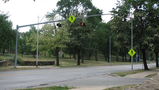 Figure 16. Photo. Unsignalized pedestrian crosswalk in Kansas City, MO. The midblock crosswalk is unmarked with pedestrian crosswalk signs (W11-2) and overhead yellow flashers, which flash continuously. The street crossed is a two-way street with two travel lanes in each direction. There are no pedestrians crossing the crosswalk in this photo.