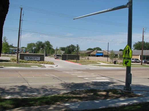 Figure 17. Photo. A different view of the crosswalk in Garland, TX. A pedestrian crosswalk in a school zone. The crosswalk has fluorescent yellow-green School Crosswalk Warning Assembly signs on each side of the road and a mast arm above the road for installation of future traffic control devices.
