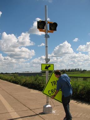Figure 55. Photo. Example of sign change. A field technician is holding a sign in preparation for inserting it into the brackets that are on the sign post that also has two circular beacons and a downward sloping arrow (W16-7P arrow plaque) below the sign.