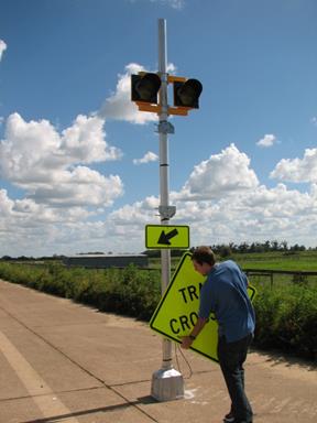 Figure 55. Photo. Example of sign change. A field technician is holding a sign in preparation for inserting it into the brackets that are on the sign post that also has two circular beacons and a downward sloping arrow (W16-7P arrow plaque) below the sign. Figure 55. Photo. Example of sign change. A field technician is holding a sign in preparation for inserting it into the brackets that are on the sign post that also has two circular beacons and a downward sloping arrow (W16-7P arrow plaque) below the sign.