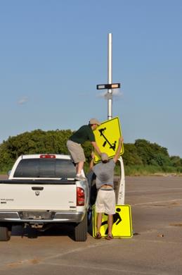 Figure 56. Photo. Another example of sign change. A field technician is standing in the back of a truck, and an other field technician is on the pavement, with both technicians holding a sign in preparation of inserting it into the brackets that are on the sign post. The sign post also has rectangular beacon light bar.