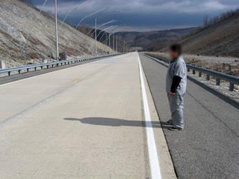 Figure 35. Photo. Pedestrian on roadway. The photo shows a straight stretch of roadway with guardrails on both sides and overhead lighting masts on the left. A man wearing gray scrubs is standing on the right side of the road and facing the road.