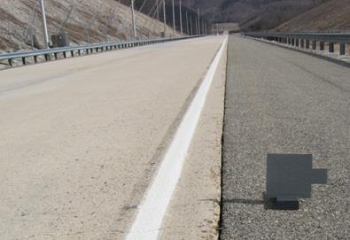Figure 36. Photo. Target on roadway. The photo shows a straight stretch of roadway with guardrails on both sides and overhead lighting masts on the left. A gray target is on the right side of the road with its tab pointing away from the road.