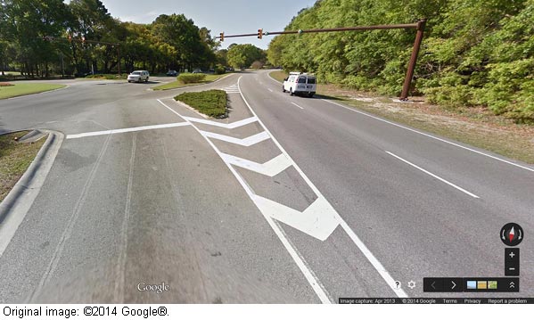 This photo shows the driver view of an approach toward continuous through lanes at a continuous green T (CGT) intersection. It shows a roadway from the perspective of a driver. The driver is approaching an intersection that separates into two branches. The right branch has two lanes that are both through lanes. There is a white van in the right-hand lane, and the two lanes are controlled by traffic lights, the left-hand of which is red and the right-hand of which is green. The left branch is a single lane that bends slightly to the left and is not controlled by a traffic light.