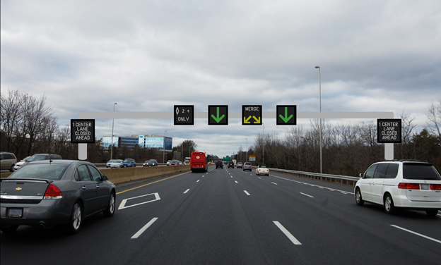 Figure 27. Photo. First picture in the incident with center lane closed scenario. This figure shows a four-lane highway with active traffic management signs over each lane and changeable message signs (CMSs) on the gantry on both the right and left. From left to right, the overhead signs show a high-occupancy vehicle lane designated by a diamond shape and the text "2+ ONLY" an open lane, a lane-closed split-merge, and an open lane. The CMSs show "1 CENTER LANE CLOSED AHEAD."