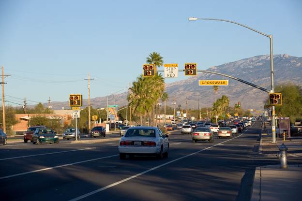 Figure 1. Photo. Example of PHB installation in Tucson, AZ. This photo shows a pedestrian hybrid beacon at a crosswalk in Tucson, AZ. The beacon is mounted on a mast arm, and the face of the beacon is composed of two red signal indications side by side and one yellow signal indication centered below the red signal indications. The signals are placed on a black back plate with a yellow border. The mast arm also has an R10-23 STOP ON RED regulatory sign next to the beacon and an illuminated pedestrian crossing sign hanging from the mast arm.