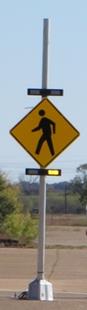 Figure 1. Photo. Study assembly containing LEDs above, below, and within the sign. This photo shows a pedestrian crossing sign supplemented with two sets of light-emitting diodes (LEDs). One set of LEDs is placed above the sign, and another is placed below the sign. In addition to the two sets of LEDs there is one set of LEDs around the border of the sign. The rectangular flashing beacon located below the sign is active, with the right-side LED illuminated.