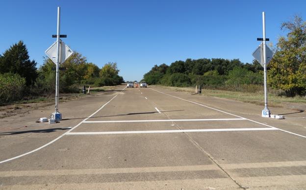 Figure 5. Photo. Back view of study site. This photo shows a back view of the study site crosswalk from the back of the crosswalk. In the background, there is a two-lane road with two parked vehicles, one per lane, with an orange work zone barrel between the vehicles. In the foreground, there is a crosswalk outlined by two solid transverse white lines on the pavement. The back side of two signs and rectangular rapid flashing beacons on each side of the road are visible.